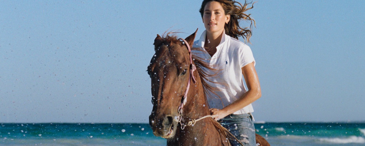 Woman in white Polo shirt & distressed jeans riding horse on beach
