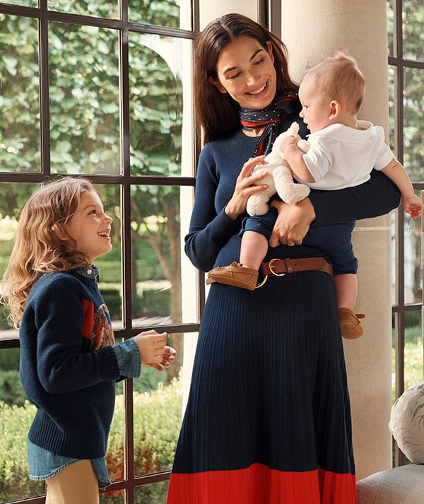 Woman in navy sweater and navy-and-orange pleated skirt holds baby.
